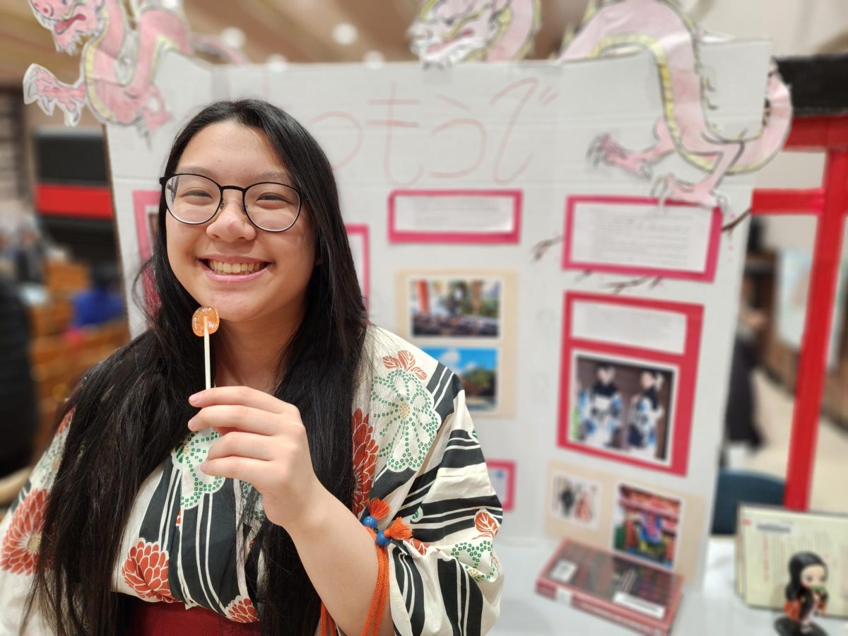 Japanese language student Carmen Yee smiling in front of a Japanese trifold, presenting at a Hatsumoude station, which means the first shrine visit of the year.
